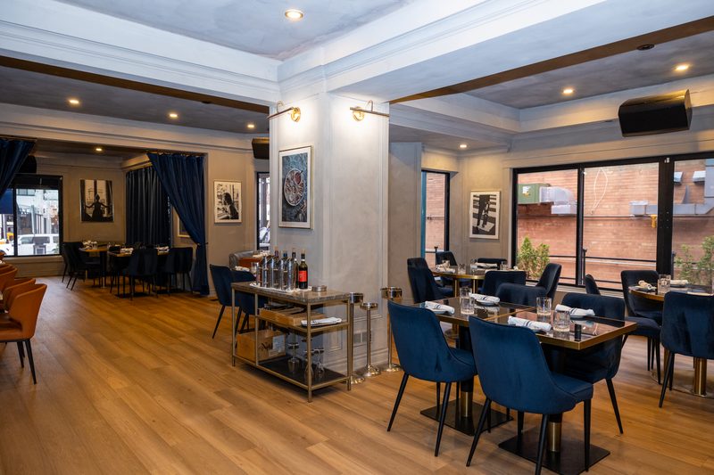 The main dining room at Beacon Social House — navy velvet chairs, brass-edged tables and floor-to-ceiling windows onto Lakeshore Road