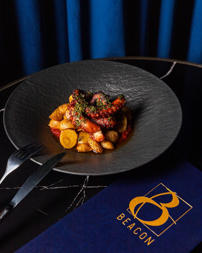 Grilled octopus with chimichurri and crushed potatoes, served on black slate beside the navy and gold Beacon menu
