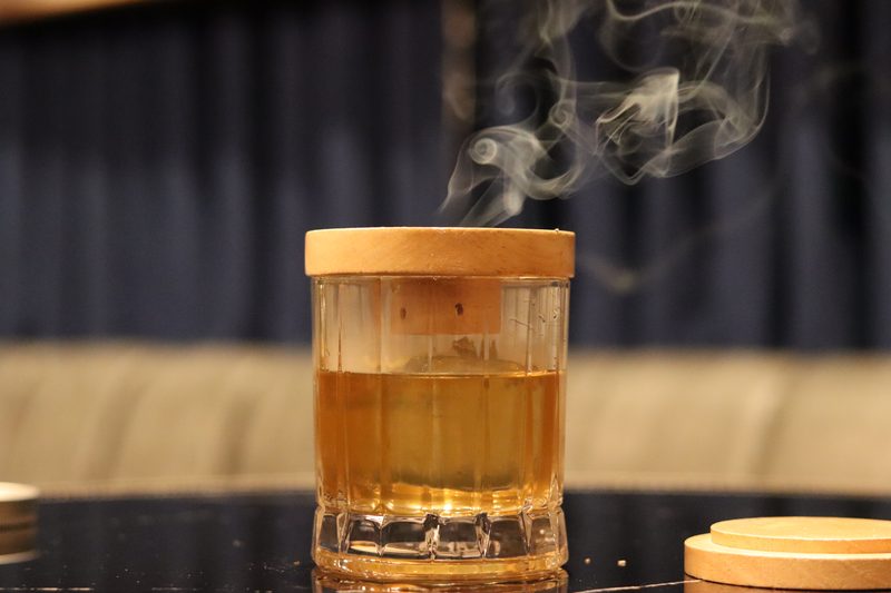 A smoked old fashioned under its wooden lid, smoke curling from the glass